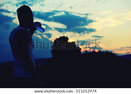 Silhouette of male jogger drinking water while enjoying the sunset with copy space area  for your text message or content, muscular build sportsman taking break after physical exercises in the park