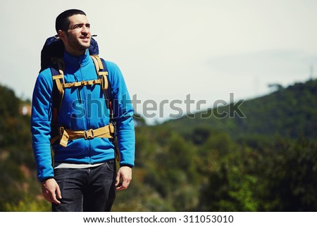 Portrait of a young tourist in mountain clothes and backpack standing on a top of mountain and enjoying scenery landscape view, hiker looking to copy space area for your text message or information
