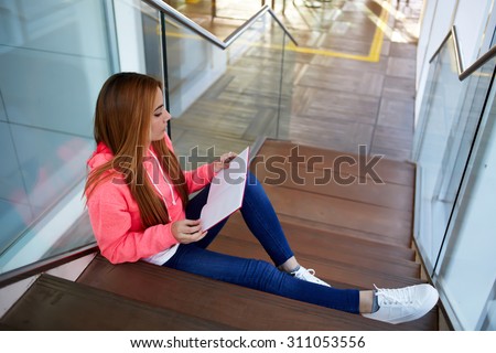 Intelligent female student reads her copybook while sitting on the wooden stairs of university hallway, stylish hipster girl preparing for classes or exams at college indoors campus, learning hard