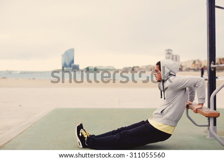 Side view portrait of strong man doing muscles exercises on training apparatus outdoors, fit man pumps biceps at seashore horizontal bar, sportsman at physical activity in cloudy cold morning, filter