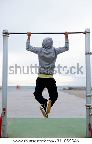 Back view portrait of  young bodybuilder in active wear doing pull ups on the horizontal bar outdoors, strong athlete doing exercise in a cloudy morning outside, fit man working out at street gym