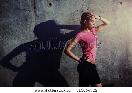 Fitness woman with beautiful figure holding hand above her head while rest after run outdoors, attractive female runner taking break after workout while standing against concrete wall with copy space