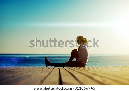 Full length silhouette of young woman runner sitting on the wooden pier against ocean while resting after an run in beautiful evening, sportswoman with good figure resting after an active run outdoors