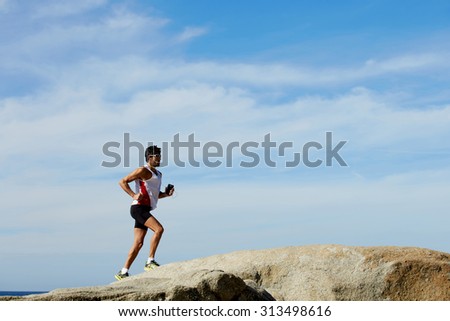 Full length portrait of mature male jogger running up the rocks against blue sky background with copy space area for text message, sports man training outdoors while listening to music in headphones