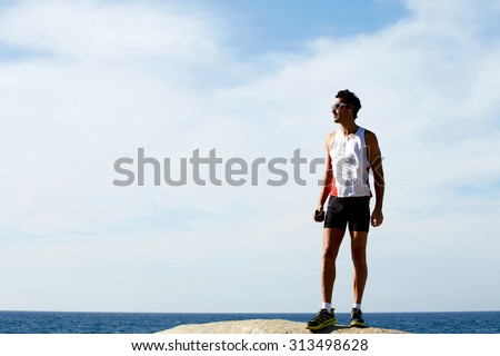 Full length portrait of mature sport man having rest after run while standing on the rock and listening to music in headphones, male runner taking break after active training outdoors in mountains