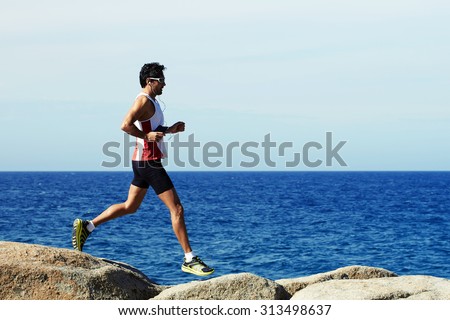 Mature man runner with perfect body jogging along the sea and jumping over big rocks while listening to music in headphones, handsome sportsman in sportswear doing an active workout outdoors
