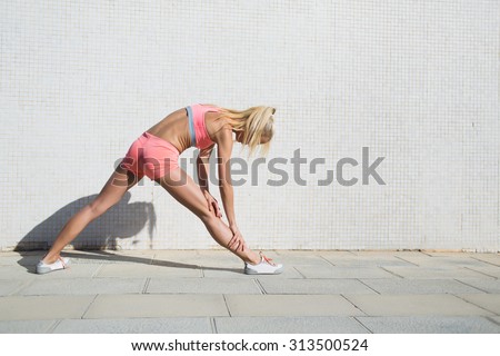 Half length portrait of young female runner stretching her muscles before begin training session in the fresh air, fit woman in sportswear stretching legs against street wall with copy space for text