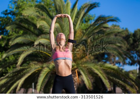 Half length portrait of athletic female with beautiful figure doing stretching arm exercise before her morning run, fit woman listening to music in headphones while working out outdoors in the park