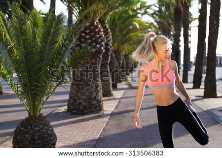 Portrait of charming female jogger stretching leg muscle while standing in palm park on running road, fit caucasian woman with beautiful figure do physical exercises outdoors, copy space area for text
