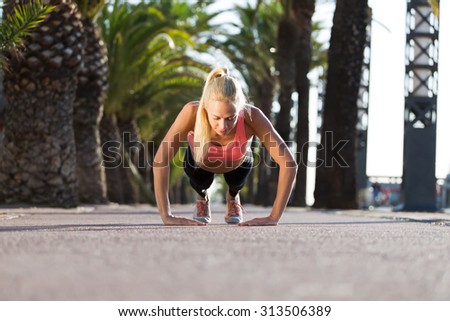 Young caucasian woman doing press ups in tropic urban setting with palm trees, athletic female runner with beautiful figure dressed in workout gear doing push-ups outdoors before begin her morning run