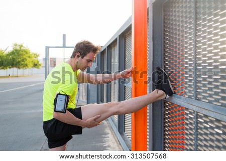 Young male runner stretching leg before jogging outdoors while listening to music in headphones on his smart phone, concentrated man doing legs exercises leaning on a metal fence at sunny day