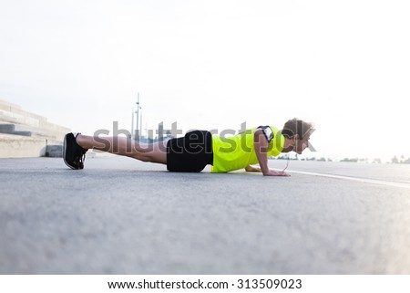 Full length view of strong build man with running armband doing push-ups on asphalt city road and listening to music with headphones,young male jogger warm up before start his workout training outside