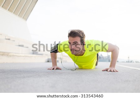 Portrait of strong build sports man with running armband doing pushes on asphalt city road and listening to music with headphones, young male jogger warm up before start his workout training outside