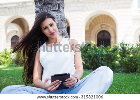 Portrait of a pretty female student reading digital e-book on touch pad at campus, beautiful latin woman holding her tablet computer while posing for the camera during rest after walking in summer day