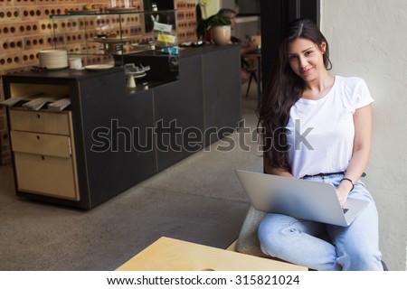 Gorgeous female freelancer using net-book for distance job while sitting in modern coffee shop,young hipster girl looking at you while working on her laptop computer and relaxing during lunch break