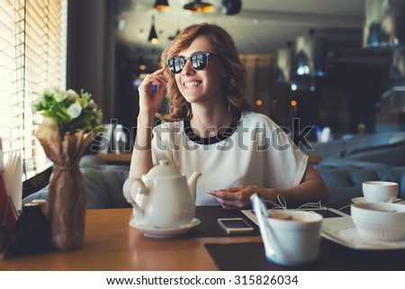 Portrait of attractive young woman smiling charmingly while looking out the window for her friends just arrived to the coffee shop where they meet,gorgeous female enjoying recreation time at breakfast