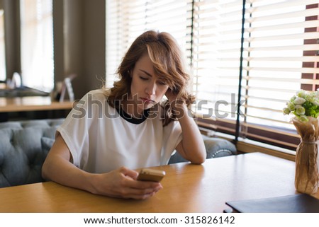 Half length portrait of lovely caucasian female reading text message on her mobile phone, young gorgeous woman holding smart phone while sitting in modern coffee shop interior near big window