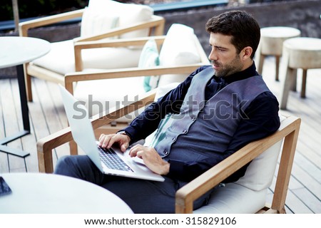 Thoughtful wealthy businessman work on-line on net-book while sits at modern restaurant terrace, young intelligent rich man connecting to wireless via laptop computer during work break in coffee shop