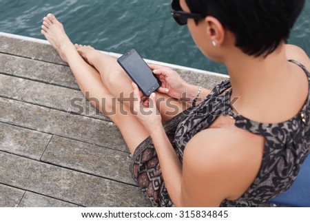 Cropped image of young woman watching video on her cell telephone while sitting on a river jetty in summer day, female using smart phone with copy space area on the screen while relaxing outside