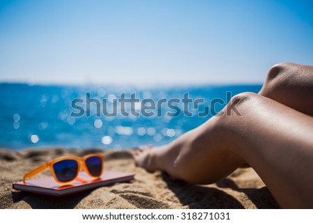 Cropped shot view of beautiful woman\'s legs on the beach near the sea and little red notepad on which lying fashionable colorful sunglasses, relaxing during summer weekend enjoying good day and sun