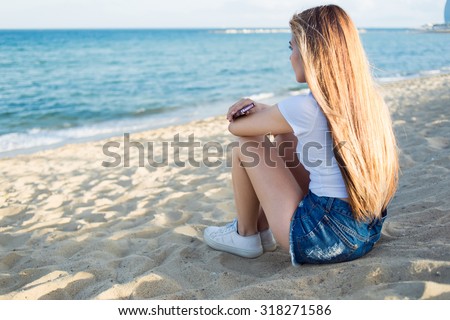 Back view of hipster girl holding cell telephone while sitting alone on the beach enjoying sea landscape in summer day,dreamy woman waiting for a call on her smart phone while relaxing after promenade