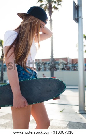 Young funky woman skater dressed in trendy clothes walking on a Miami street during summer holiday with her skateboard with copy space for you brand, stylish hipster girl strolling with long board