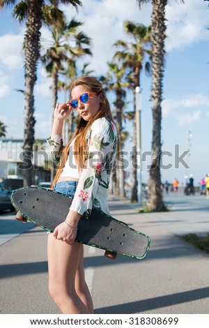 Fashionable woman dressed in trendy clothes showing lips kiss while posing with  long board against sky and palm trees,stylish female skateboarder walking on promenade with penny board in sunny day