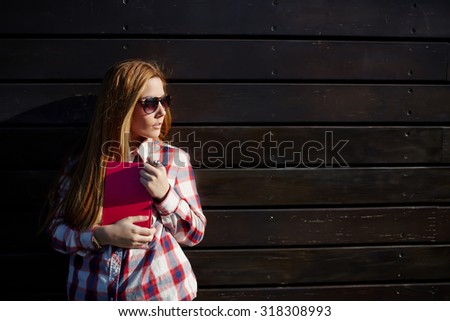 Half length portrait of young university student holding pink book in the hands with copy space area for text, charming female in sunglasses standing on wooden wall background with a notebook in hands