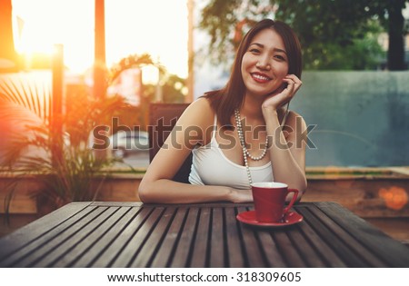 Portrait of a smiling beautiful asian woman enjoying free time while sitting in coffee shop exterior terrace with beautiful sunset on background, wonderful young female posing for the camera in summer