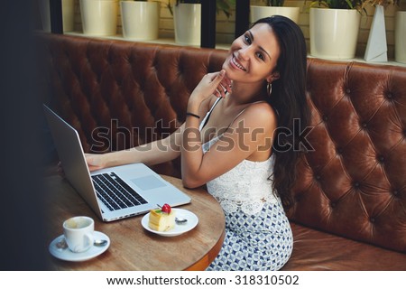 Smiling happy latin woman enjoying a cup of coffee and dessert while working on her laptop computer during lunch break, successful freelancer girl using net-book while sitting in modern coffee shop