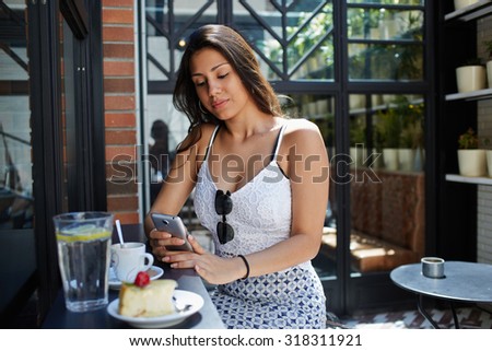 Young female student use mobile Internet on smart phone to correspond with her friends in chat,modern arabic women typing text message on cellphone while breakfast at sidewalk cafe in sunny summer day