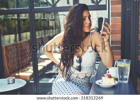 Young well dressed woman with a perfect figure looking to her smart phone screen while breakfast in restaurant terrace,attractive latin female browsing internet on her touchscreen phone at coffee shop