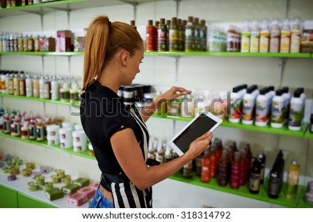 Young beautiful female consultant checks the quality of cosmetic product verifying information on digital tablet, woman entrepreneur puts pharmaceutical production on trade shelves in her drugstore