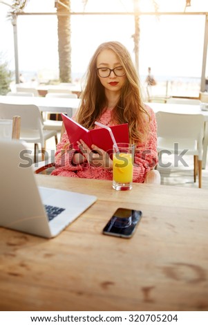 Attractive creative freelancer read notebook while sitting at wooden table with laptop laptop computer and cellphone, young female woman having breakfast on terrace in modern coffee shop, flare sun