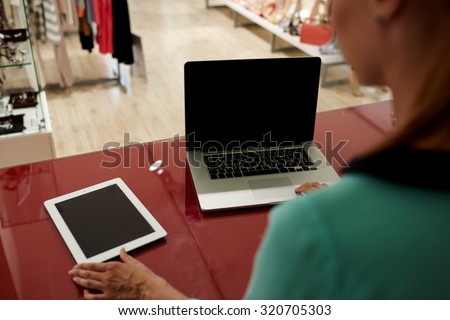 Seller woman working in the shop on net-book and digital tablet with copy space area black screen for text message or advertising, touch pad wireless device and open laptop lying on cash store table