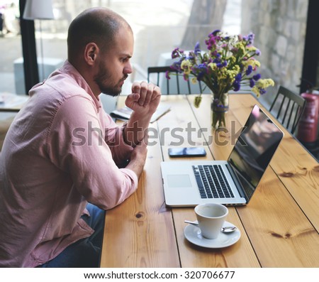 Thoughtful businessman work on net-book while sitting at wooden table in modern coffee shop interior, student reading text or book in cafe, male freelancer connecting to wireless via laptop computer