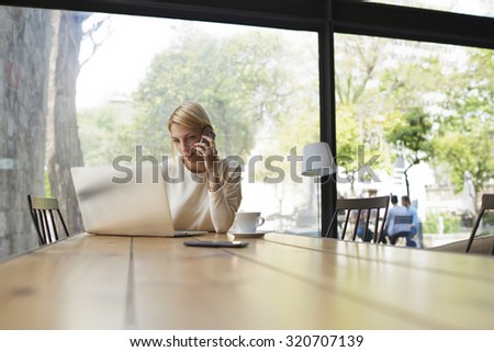 Gorgeous female speaking on mobile phone with smile while sitting at wooden table with open laptop computer, young student or businesswoman at work break with net-book in modern coffee shop interior
