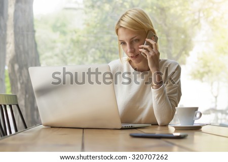 Portrait of experienced young businesswoman at work break keyboarding text on laptop computer while talking on smart phone, female person busy working in modern office interior or coffee shop, flare