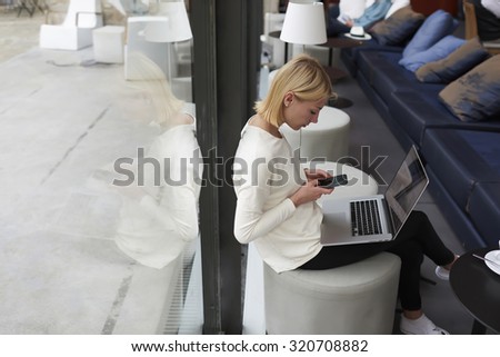 Female student typing text message on smart phone sitting at library with laptop computer, freelancer woman working on net-book in modern coffee shop interior, young businesswoman at coffee break