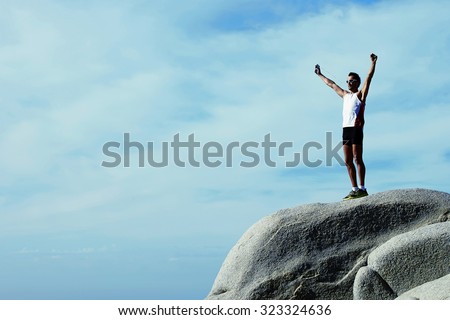 Male runner relaxing after training while standing on stone rock with hands raised against sky background with copy space area for your advertising, fit men celebrating achievement with hands raised