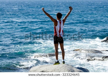 Full length portrait of fit men feeling free while standing on a stone rock against blue sea, athletic male with raised hands celebrating achievement,sportsman enjoying rest after workout on the beach