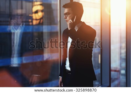 Portrait of successful businessman talking on mobile phone while standing against window in hallway of modern office interior, young confident man having cell telephone conversation during work break