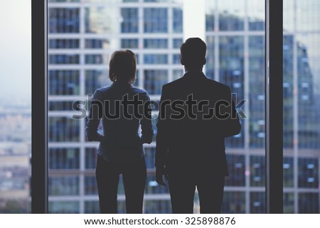 Rear view silhouettes of two business partners looking thoughtfully out of a office window in situation of bankruptcy,team of businesspeople in fear or risk watching cityscape from skyscraper interior