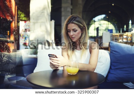 Portrait of a cute blonde female read something on her smart phone while sitting in modern coffee shop, beautiful young hipster girl using cell telephone while enjoying her free time in expensive cafe