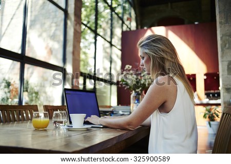 Portrait of a young female freelancer using laptop computer for distance job while sitting in modern coffee shop interior, smart blonde woman working on net-book during morning breakfast in cafe bar