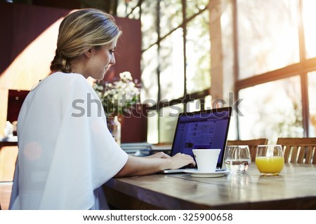 Rear view of a successful pretty female freelancer using net-book for distance job while sitting in modern coffee shop interior, young Sweden woman working on laptop computer during morning breakfast