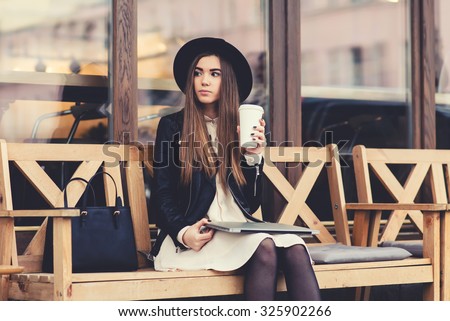 Portrait of a glamorous young woman holding on her knees portable laptop computer while sitting on a wooden bench, stylish female drinking coffee while relaxing after work on net-book during free time