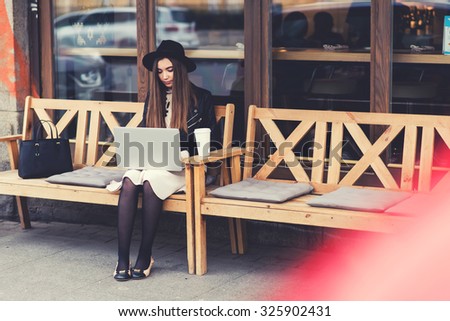Charming young woman with trendy look work on portable laptop computer during coffee break outdoors,gorgeous female freelancer using net-book for remote job while sitting on the bench in sidewalk cafe