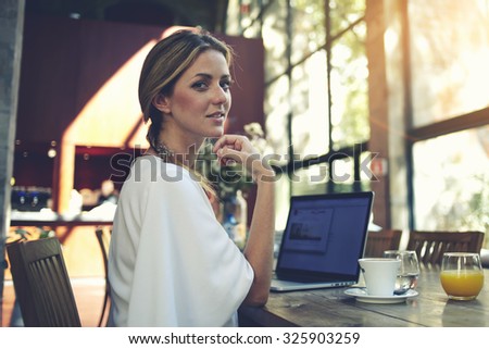 Portrait of charming smiling businesswomen posing while resting after work on her portable laptop computer during coffee break, young pretty female using net-book while sitting in modern cafe interior