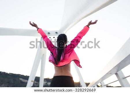 Back view of fit woman with slender body standing with raised hands felling freedom while rest after workout, young happy female enjoying good evening and weather after training in the fresh air